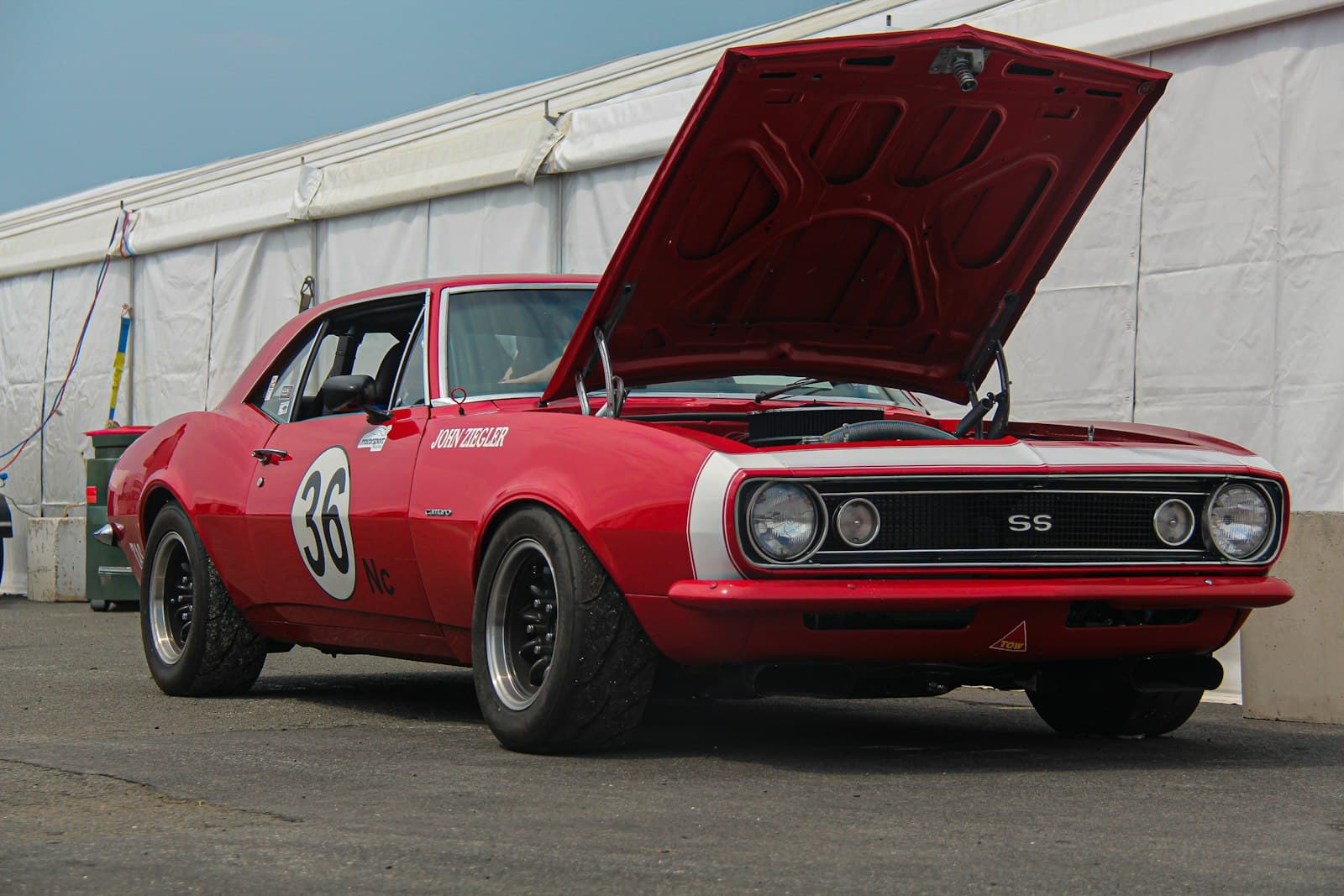 A red 1967 Camaro SS race car with a door number, hood up in the paddock